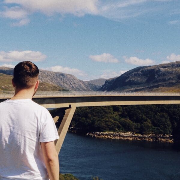 A man stands with his back to us looking at the Kylesku Bridge in the distance