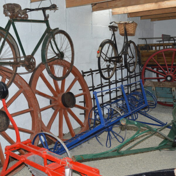 A collection of old farming equipment, including cart wheels and a plough, along with two bikes and a full horse drawn cart inside the Croft Museum.